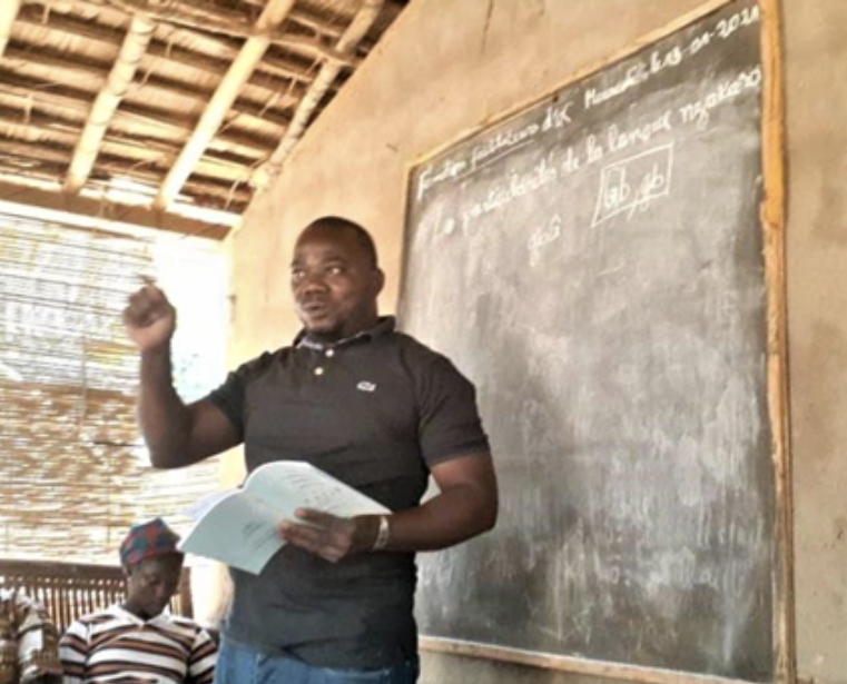 A man stands in front of a chalkboard, holding a book in one hand and gesturing with the other